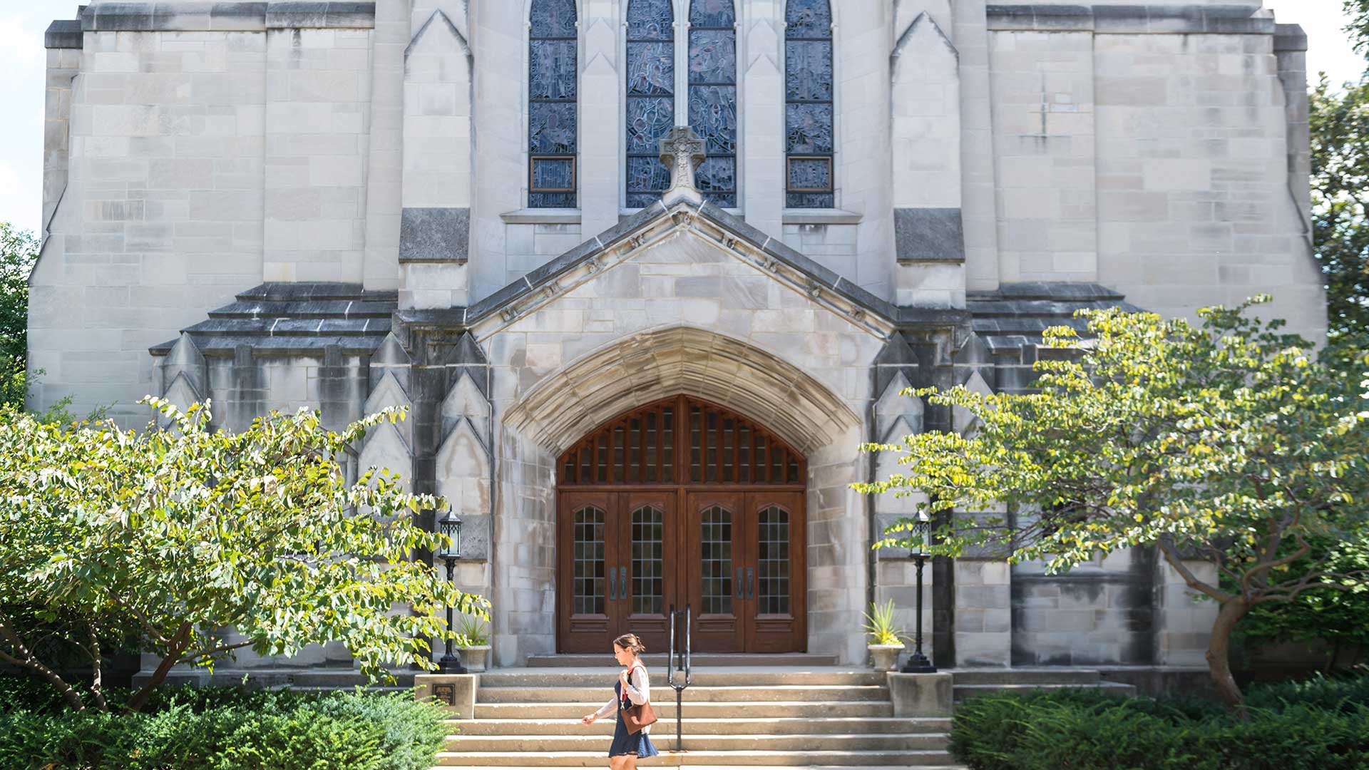 A person walks past the grand stone entrance of a historic church in Oak Park, Illinois, featuring arched doors, stained-glass windows, and lush greenery framing the scene.