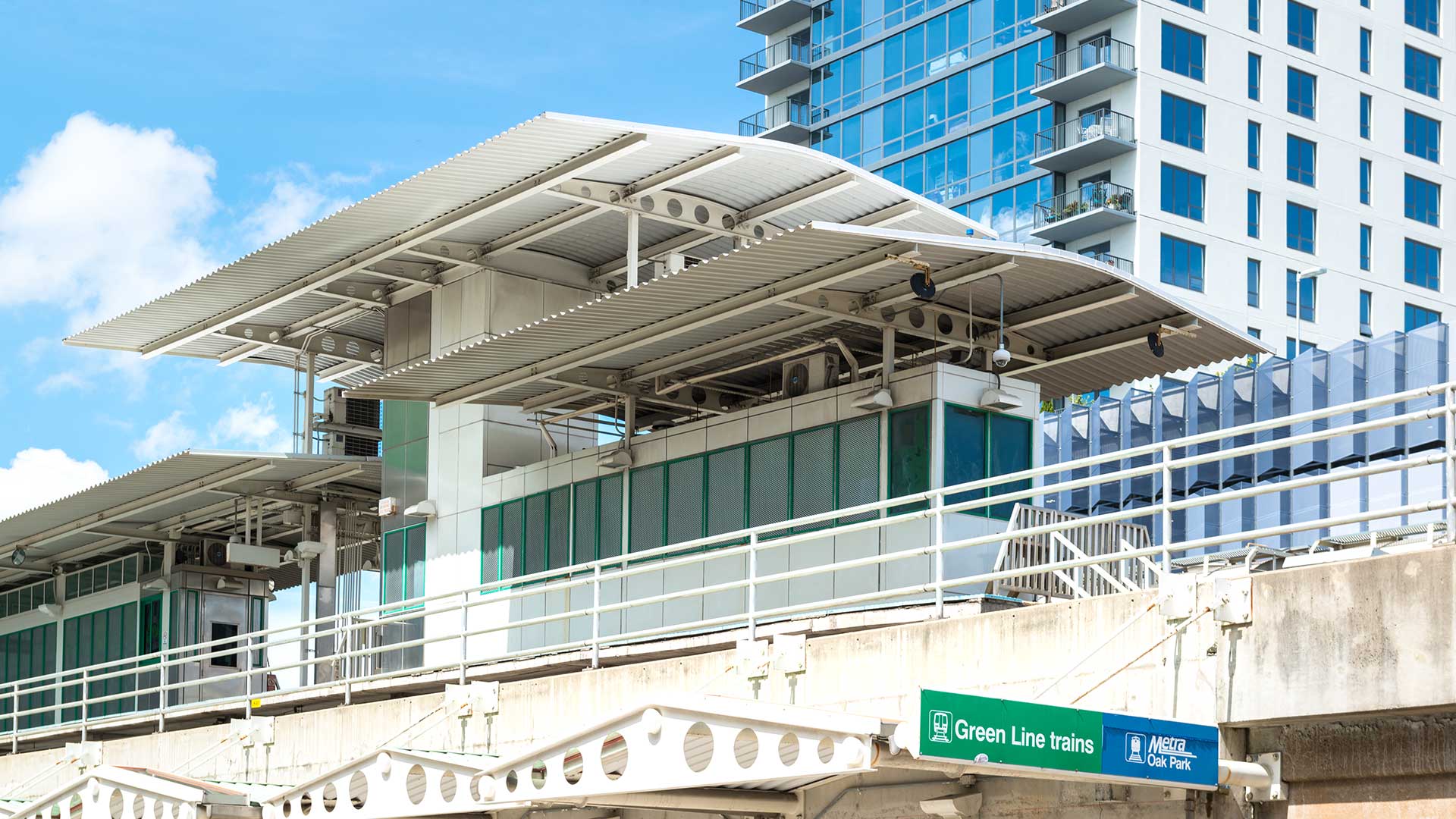 Modern elevated train station in Oak Park, Illinois, showing signage for the CTA Green Line and Metra Oak Park stop against a backdrop of contemporary glass buildings and blue sky.