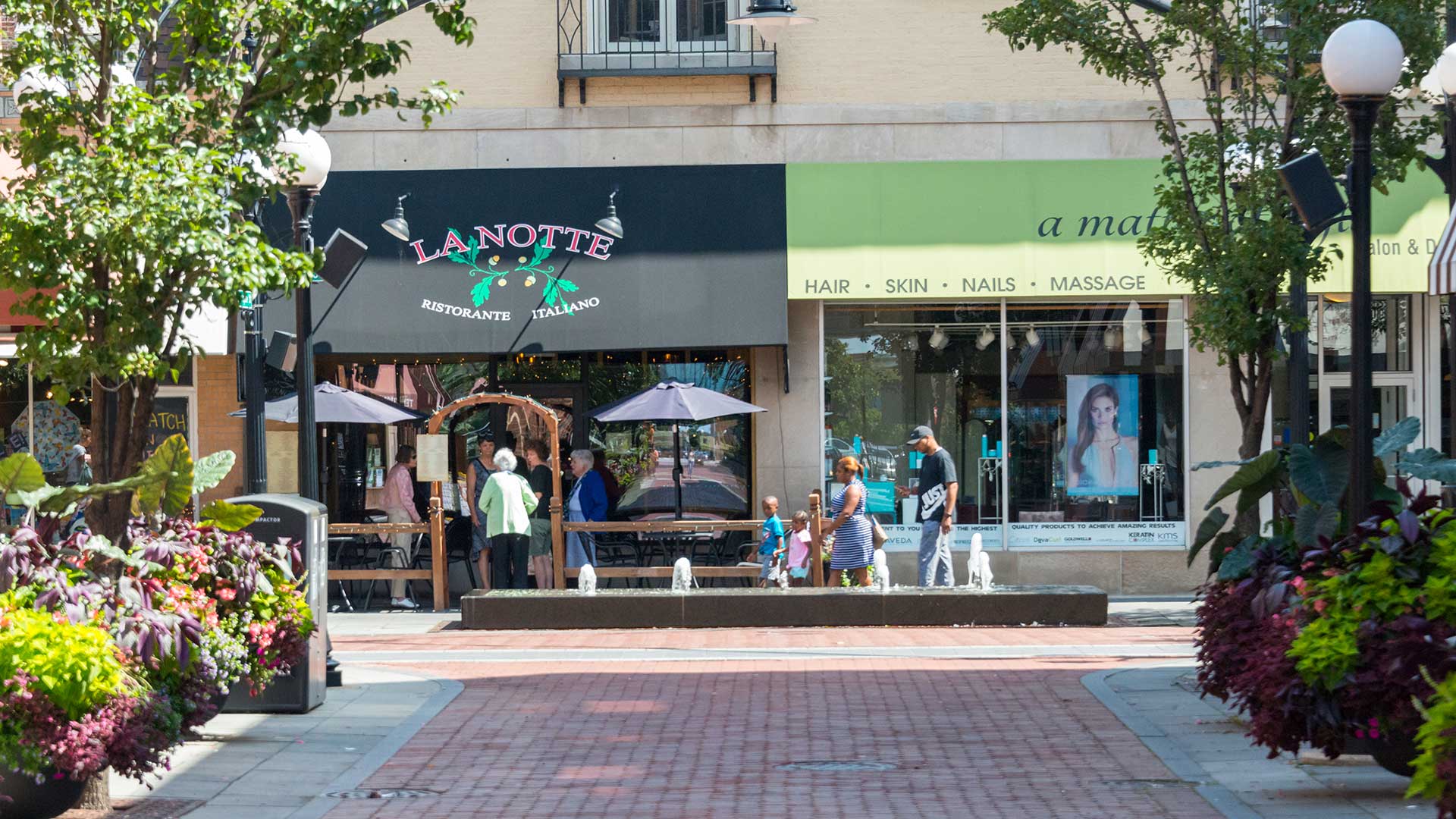 People stroll and gather near shops and a restaurant patio in downtown Oak Park, Illinois, surrounded by planters and fountains on a sunny day.