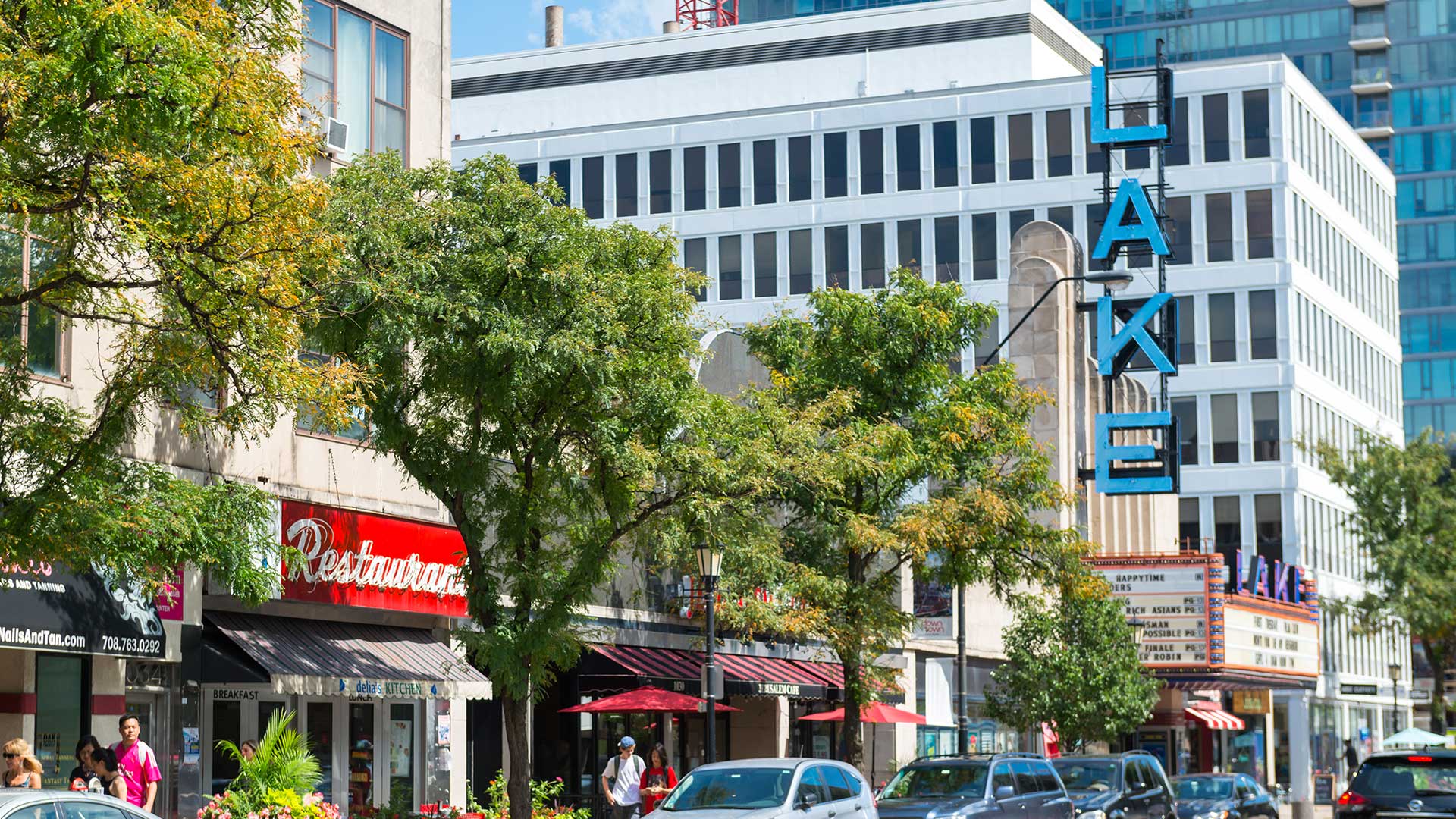 Street view of downtown Oak Park, Illinois, showing restaurants, storefronts, and the iconic Lake Theatre sign on a sunny day with trees lining the sidewalk.
