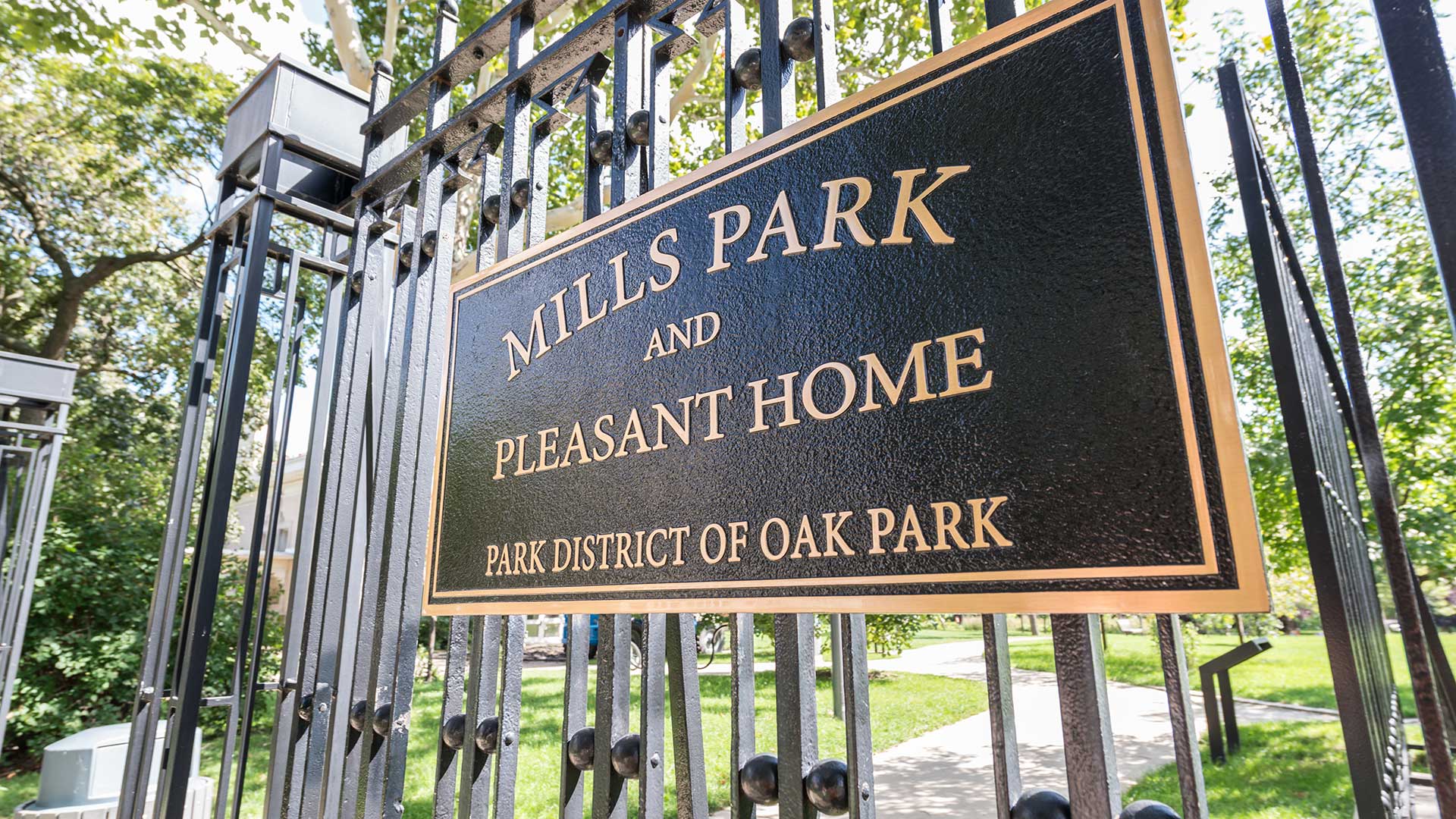Close-up of the Mills Park and Pleasant Home entrance sign in Oak Park, Illinois, with black iron fencing, green trees, and sunlight in the background.
