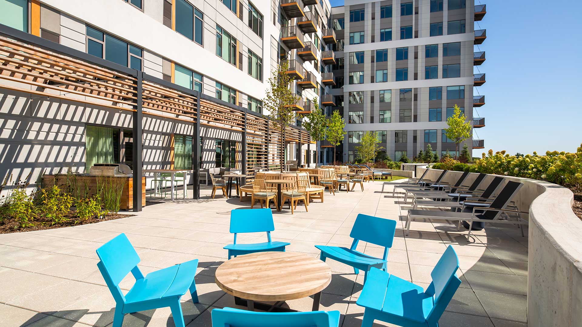 Outdoor lounge terrace at Eleven33 Apartments in Oak Park with bright blue chairs, dining tables, and sun deck seating surrounded by greenery.