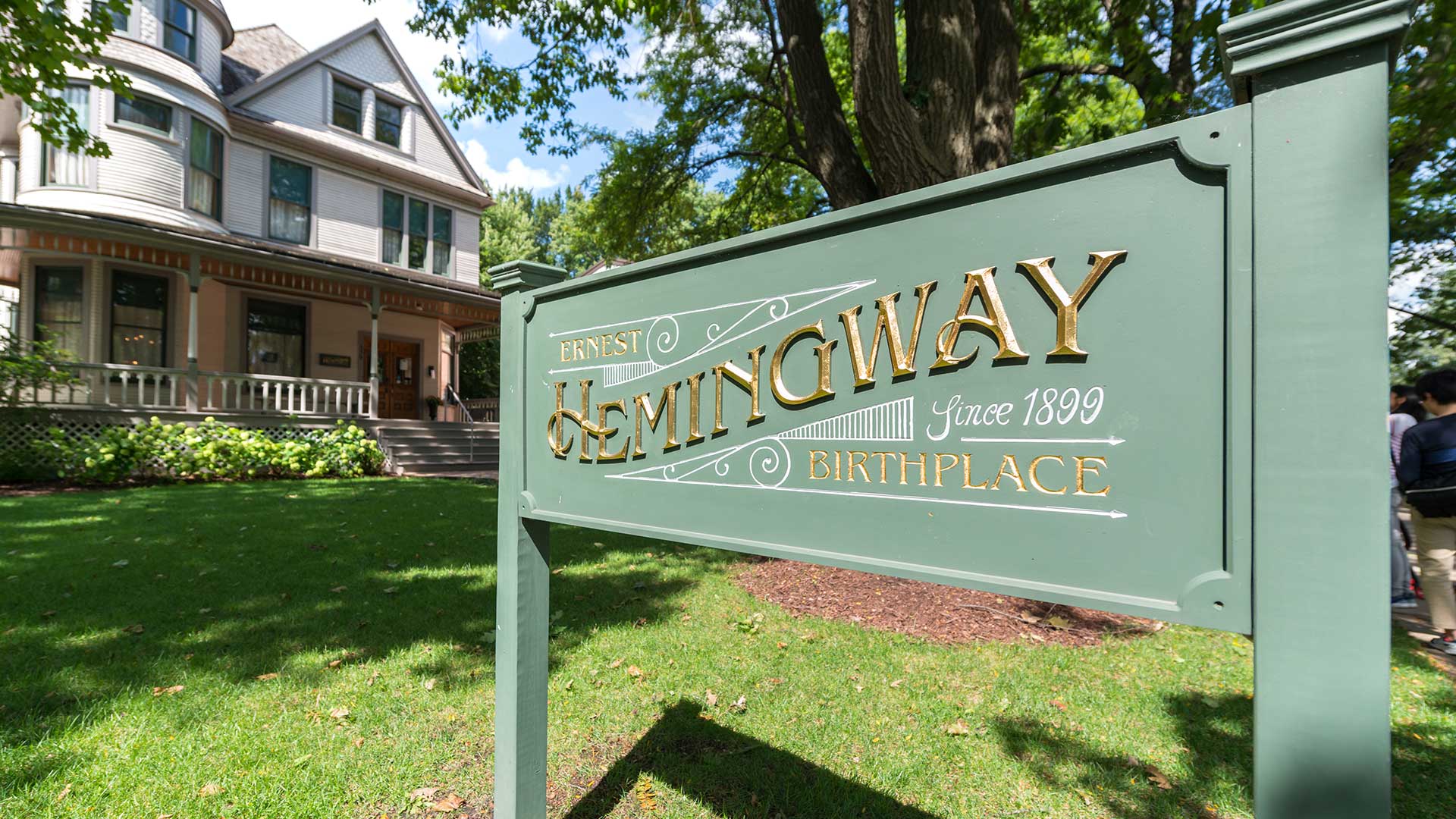 A close-up of the Ernest Hemingway Birthplace sign in Oak Park, Illinois, with the historic Victorian-style home visible in the background surrounded by trees and greenery.